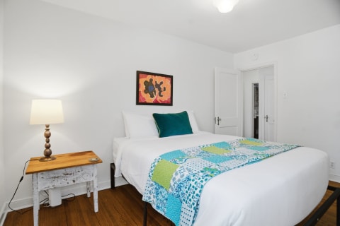A serene bedroom with a white bed, colorful quilt, a wooden nightstand with a lamp, and a piece of abstract art on the wall.