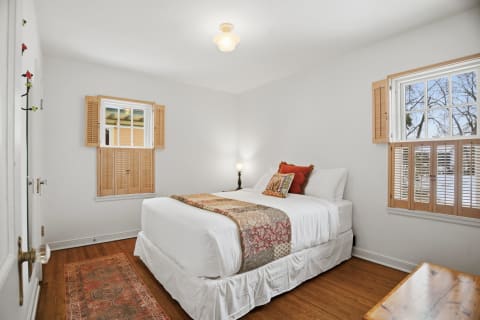 A cozy bedroom with a bed dressed in white linens and colorful pillows, warm wood shutters on the windows, and a wooden bedside table.
