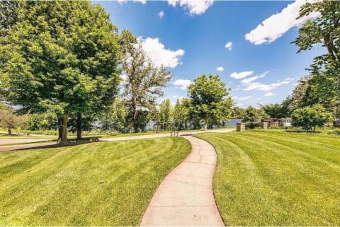 A path winding through a green lawn towards a lake, surrounded by trees and a clear sky.