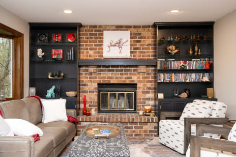A living room featuring a brick fireplace, black shelves with decorative items, a beige couch, and an armchair.