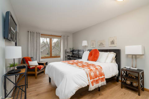 Cozy bedroom featuring a bed with white linens and orange accents, a chair, and a window view.