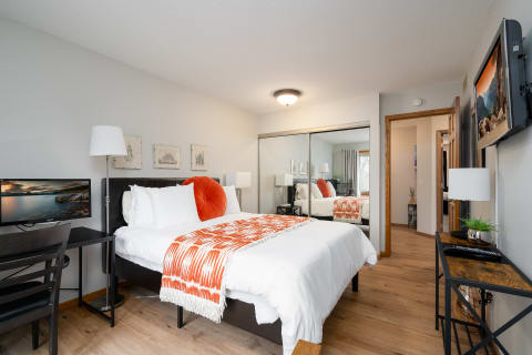 A cozy bedroom featuring a bed with an orange throw pillow, a desk with a monitor, and stylish decor.