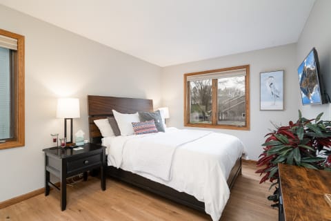 A well-decorated bedroom featuring a wooden bed, nightstand with a lamp, and a window displaying greenery outside.