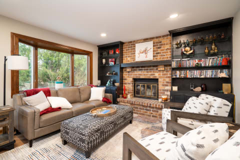Interior of a cozy living room featuring a leather sofa, a patterned ottoman, and a brick fireplace.