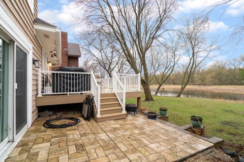 A backyard featuring a wooden deck, patio stones, and several trees near a body of water.