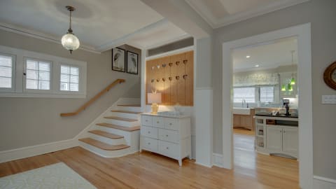 A stylish entryway with a staircase, white dresser, and a glimpse of the kitchen in the background.