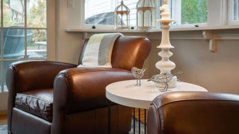 A cozy reading nook with a leather armchair, side table, wire bird sculptures, and a window showing outdoor greenery.