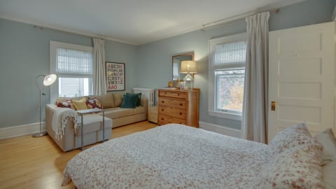 A bright bedroom with a sectional sofa, dresser, and floral bedding, showcasing autumn scenery through the window.