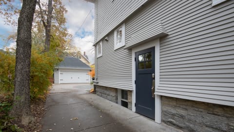 A contemporary house with gray siding, featuring a blue door and stone foundation, surrounded by trees and a driveway leading to a garage.
