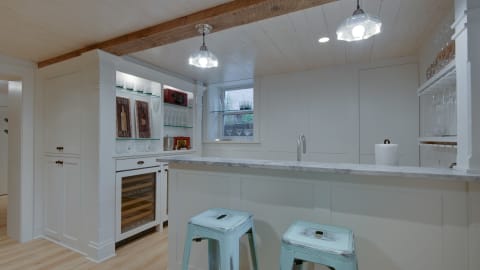 A modern kitchen with marble countertops and rustic wooden beams, featuring blue metal stools and a wine refrigerator.