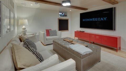 A cozy living room featuring a white sectional sofa, a tufted leather ottoman, a red media console, and a large TV, designed for entertainment.