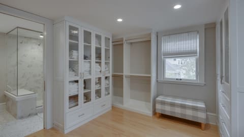 Spacious walk-in closet with glass-front cabinets and a modern marble shower in view.