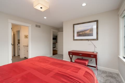 A modern bedroom with a red bedspread, a framed coastal photograph, and a red desk.