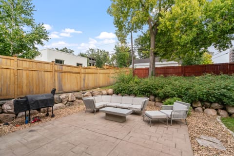 Outdoor patio with gray sectional sofa, coffee table, and a grill, bordered by a wooden fence and greenery.