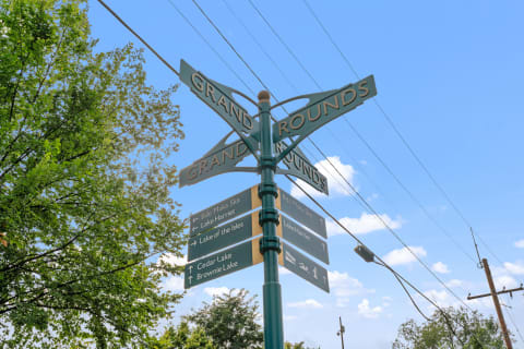 A decorative signpost labeled 'Grand Rounds' with directional arrows pointing to various local lakes against a blue sky.