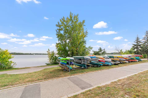 Stacked kayaks and canoes near a calm lake against a bright sky.