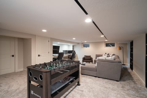 A modern basement featuring a foosball table, a gray sectional couch, and a workspace with computers.