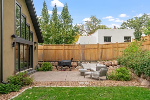 Modern backyard patio with seating and grill, surrounded by green grass and trees.