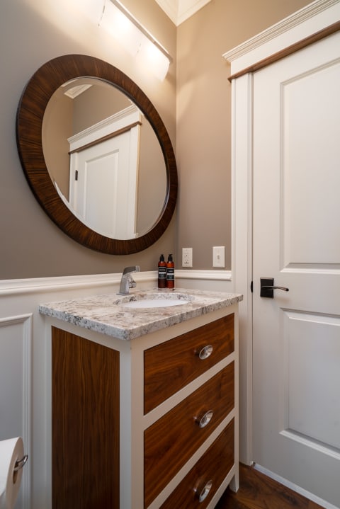 A modern bathroom corner showing a wooden vanity with a sink and mirror against taupe walls.