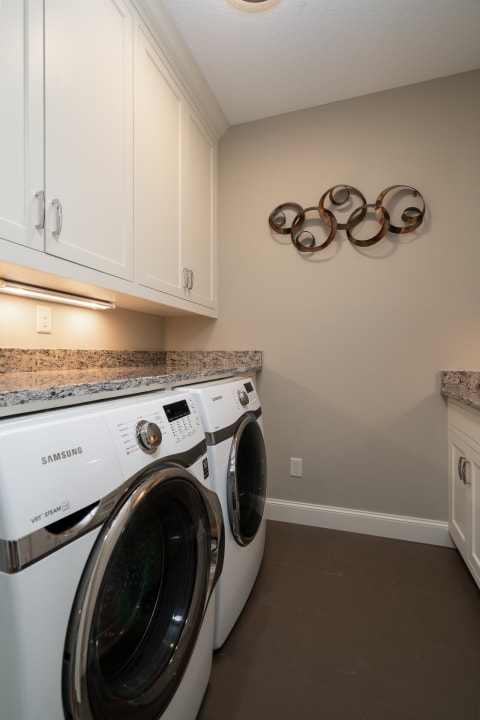 A modern laundry room with white Samsung washing machines, granite countertop, and decorative wall art.