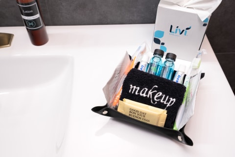 A black leather basket filled with personal care items including a makeup towel, toiletries, and a pack saying 'GOOD DAY BONJOUR BUENOS DIAS' on a bathroom counter.
