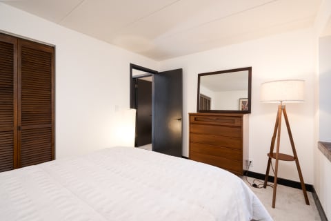 Modern bedroom with a white bedspread, wooden dresser and mirror, and a tripod floor lamp.