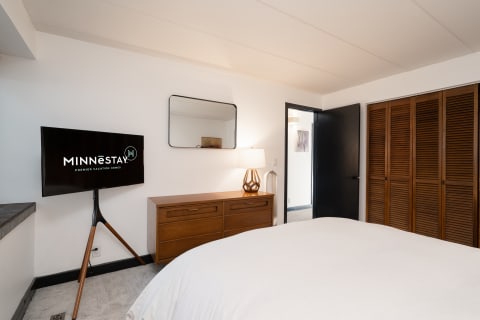 A cozy bedroom with a white bedspread, wooden dresser, and flat-screen TV.