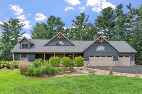 A single-story blue-gray house with wooden accents, featuring a porch and two-car garage, set in a green landscape.