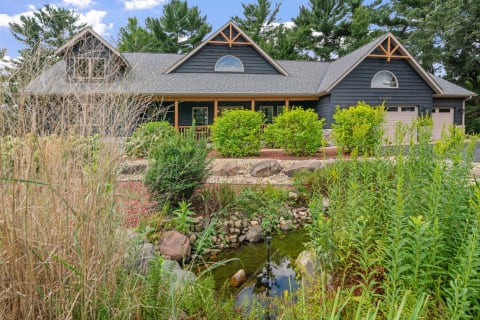 Modern dark blue house with a front porch surrounded by greenery and a pond.