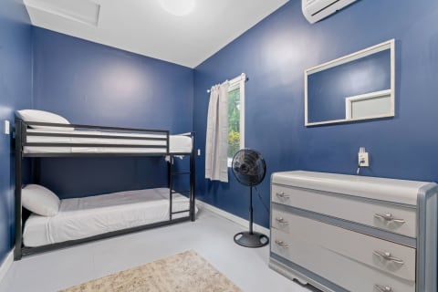 An inviting bedroom with blue walls, featuring a bunk bed, dresser, fan, and natural light from a window.