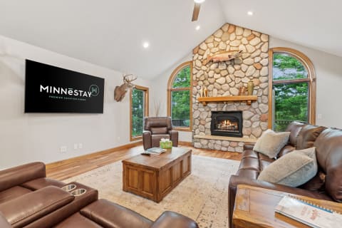 A warm living room with a stone fireplace, leather seating, and large windows overlooking greenery.
