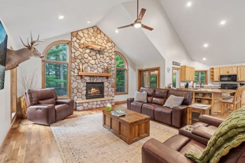 A cozy living room featuring a stone fireplace, leather sofas, and a view of trees outside.