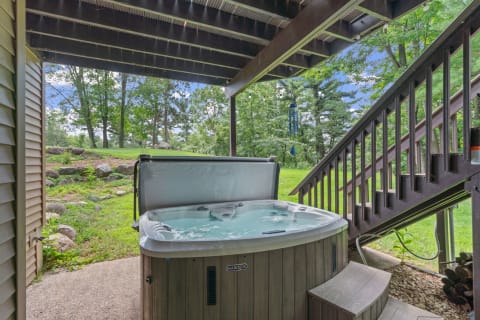 An outdoor hot tub beneath a wooden overhang with greenery in the background.