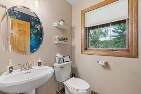 A modern bathroom with a round mirror, pedestal sink, and window overlooking greenery.