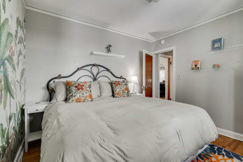 A cozy bedroom featuring light gray walls, a plush bed with floral pillows, and a tropical mural.