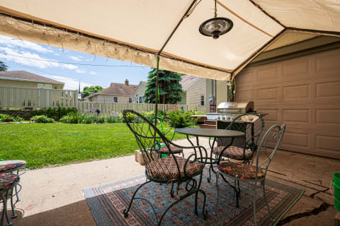 Outdoor seating area with a round table and chairs under a fabric canopy, surrounded by a garden.