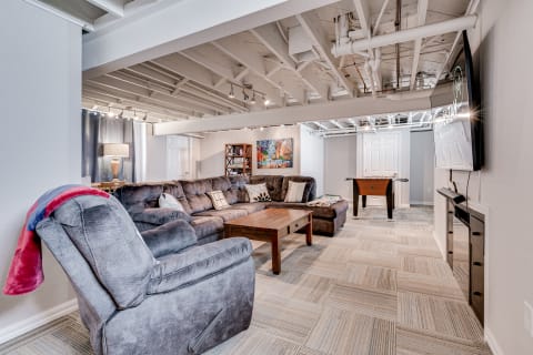 A cozy basement living room featuring a sectional sofa, a wooden coffee table, a foosball table, and colorful artwork.