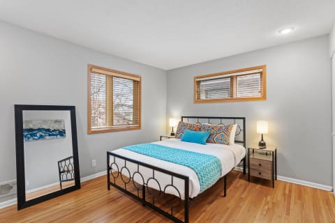 A cozy bedroom with a colorful bedspread, a mirror, and natural light coming in through the windows.