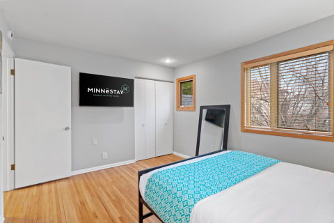 Cozy bedroom with wooden flooring, a large mirror, and a TV displaying MINNĒSTAY logo.