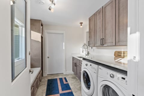 A modern laundry room showcasing white appliances, wooden cabinets, and a blue geometric rug.