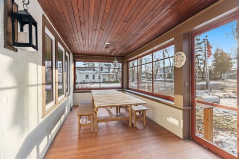 A sunroom with wooden ceiling, large windows, and a wooden dining table, overlooking a snow-covered street.