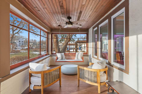 A bright sun porch with a ceiling fan, swing seat, and comfortable wooden seating overlooking a tranquil outdoor view.