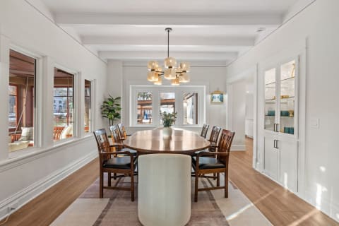 A modern dining room featuring a long wooden table with chairs, illuminated by a contemporary chandelier and large windows.