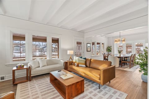 An elegant living area featuring a leather sofa, a cozy light couch, a wooden coffee table, and spacious windows, with a glimpse of a dining area.