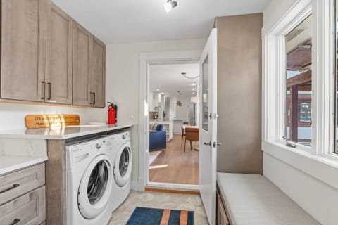 Modern laundry room featuring front-loading washing machines, wooden cabinetry, and an open doorway leading to a cozy living area.