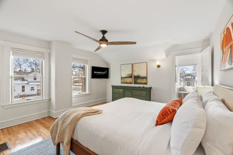 A bright bedroom with a bed, green dresser, and large windows showing a neighborhood view.