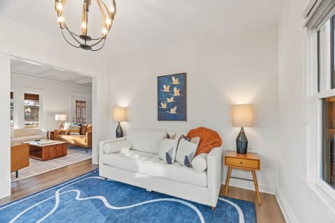 A stylish living room featuring a light sofa, orange throw blanket, blue rug, and decorative lamp, with a framed bird artwork on the wall.