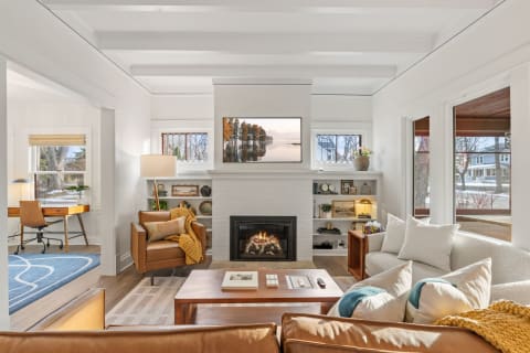 A spacious living room with a fireplace, comfortable seating, and a workspace area, illuminated by sunlight.