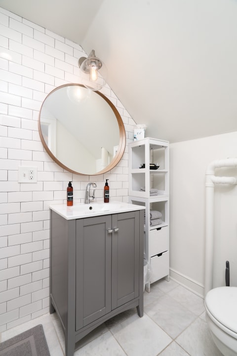 A modern bathroom with a gray vanity, round wooden mirror, and white shelving unit filled with towels.