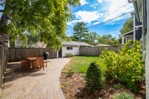 A serene backyard with a brick pathway, wooden dining set, and lush greenery.